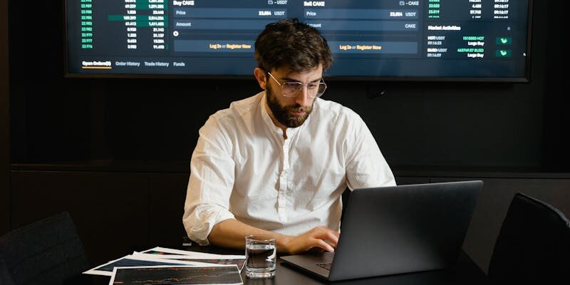 A focused man analyzing stock market trends on a laptop in an office setting with charts displayed.